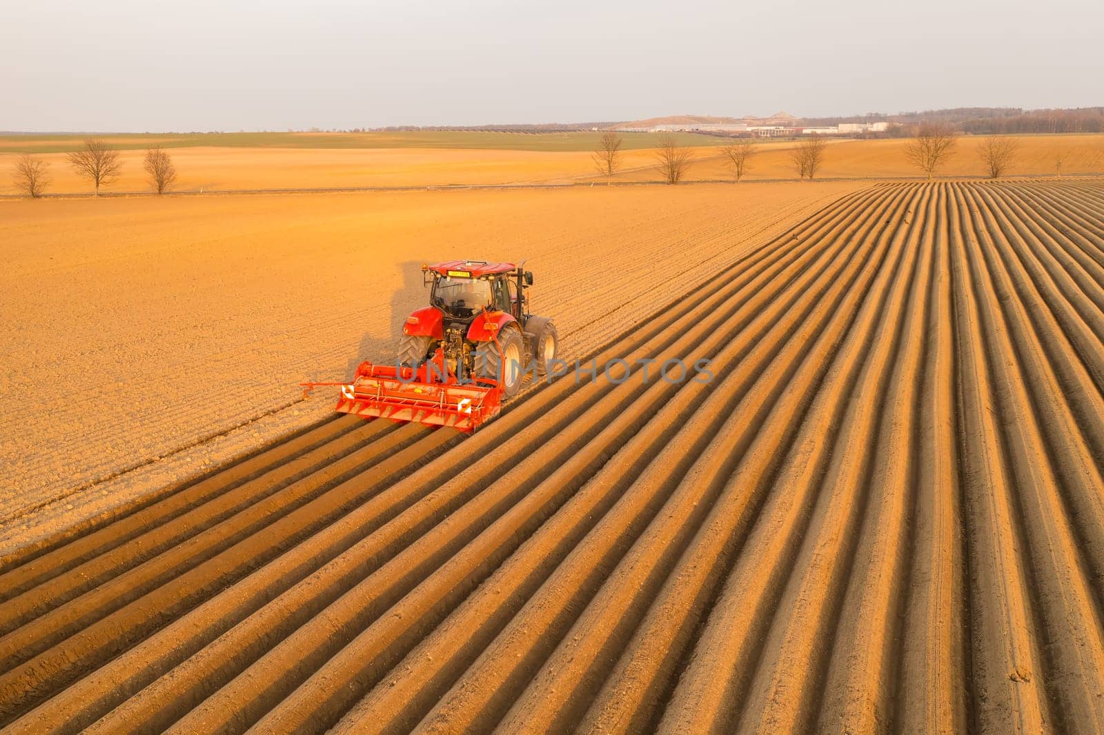 PRAGUE , CZECH REPUBLIC - MARCH 18 2022: Heavy tractor uses plow to make beds for planting potatoes in agricultural field. Powerful machine operates at farm on sunny day aerial view