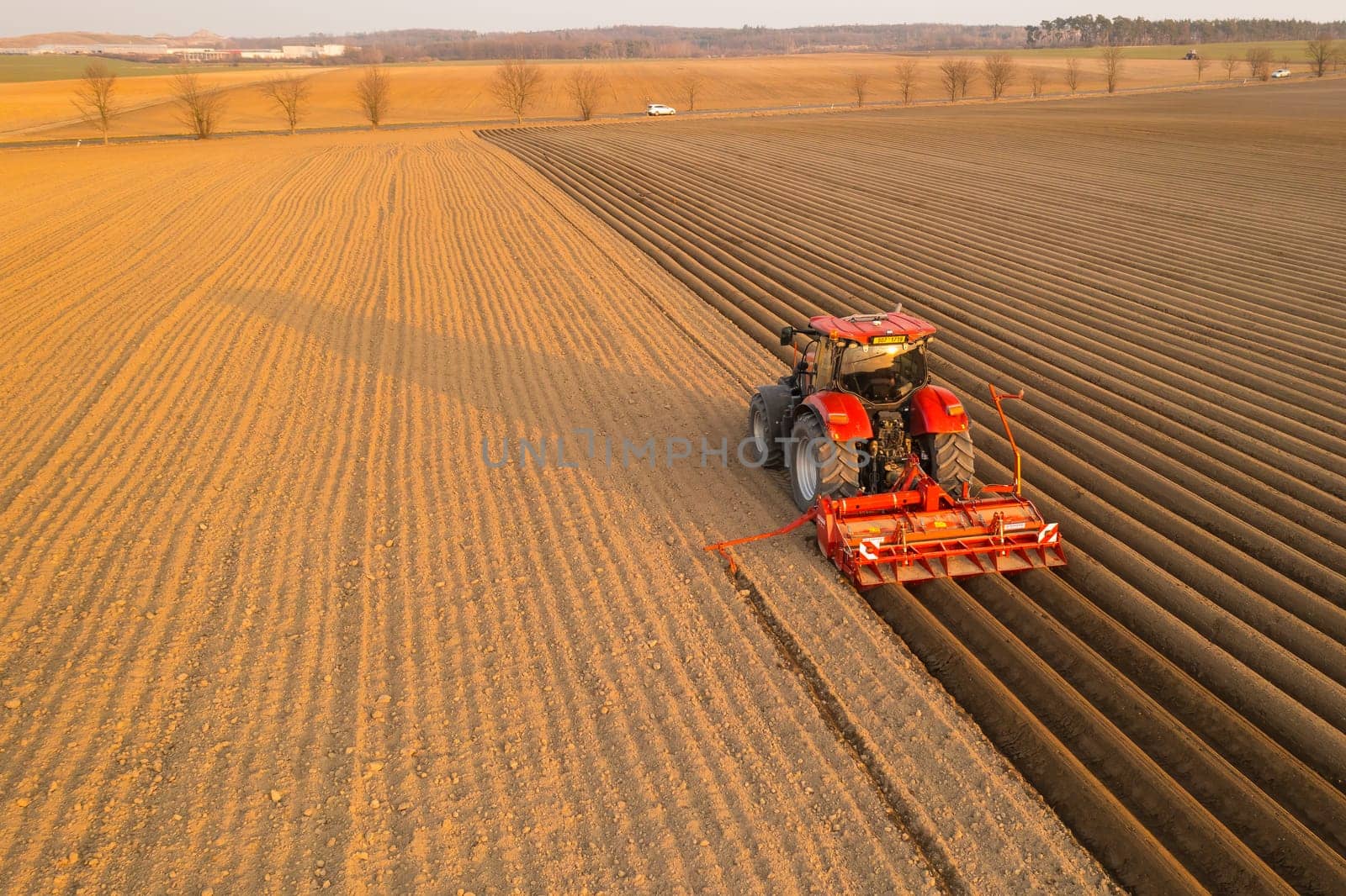 PRAGUE , CZECH REPUBLIC - MARCH 18 2022: Tractor drives at sunset leaving beds for planting vegetable seedlings in cultivated soil. Powerful machine operates on field at farm aerial view
