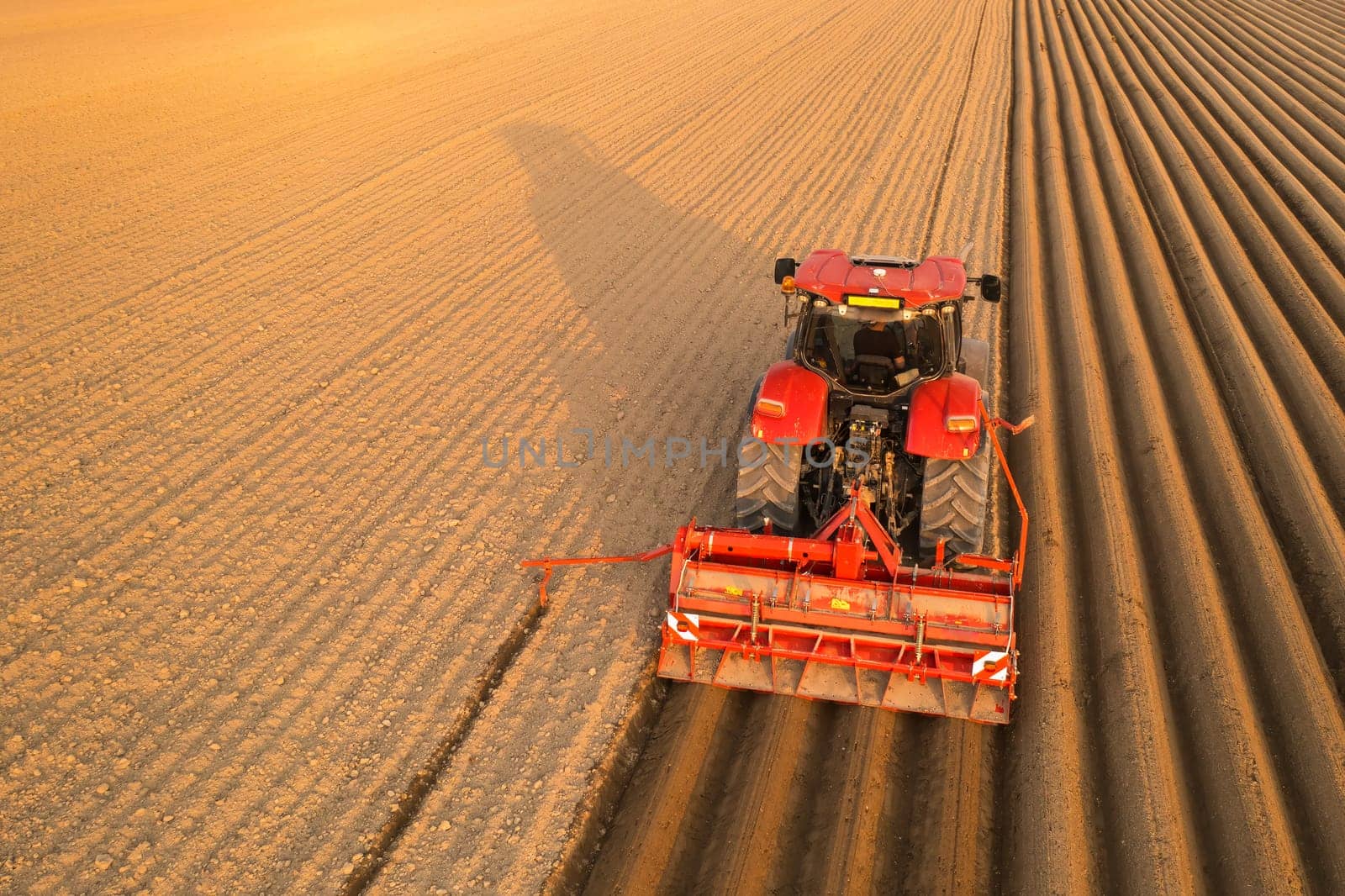Tractor drags plow for leaving long furrows behind in soil on sunny day. Agricultural transportation works in endless field at sunset aerial view