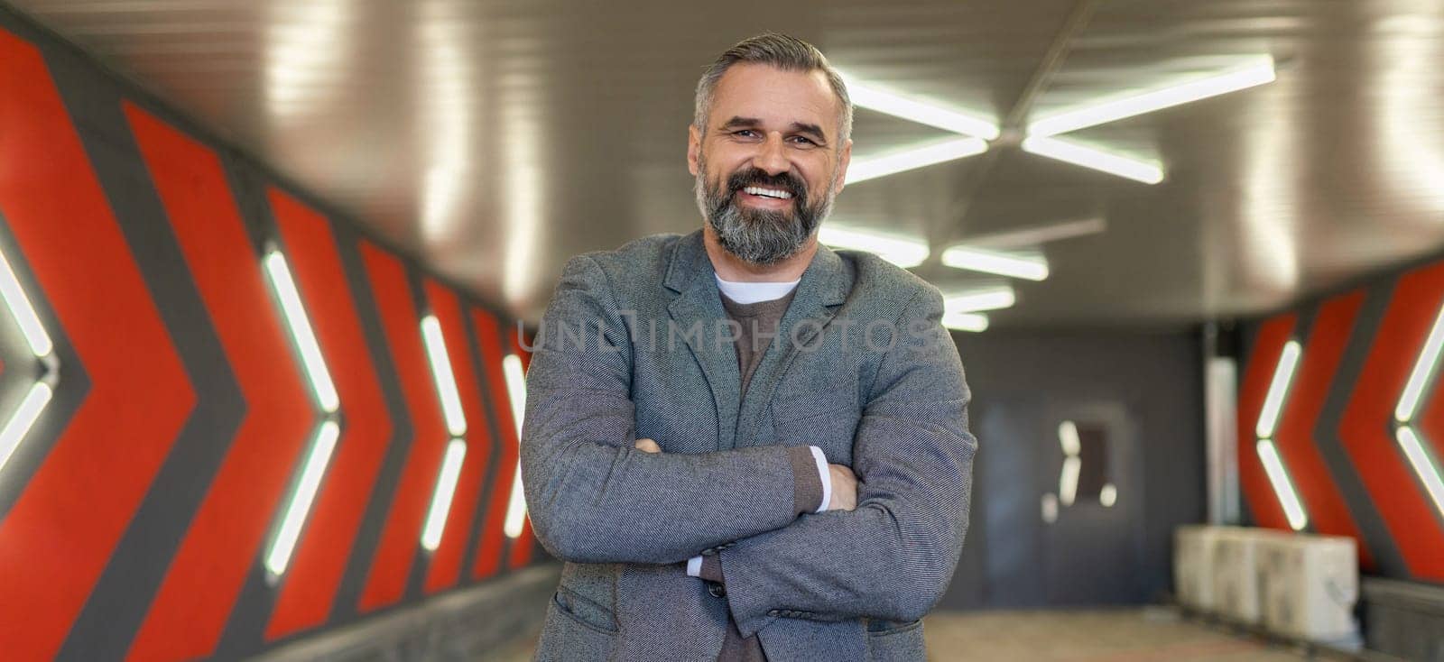 A gray-haired fifty-year-old man businessman in an underground parking lot stands with his arms folded across his chest.