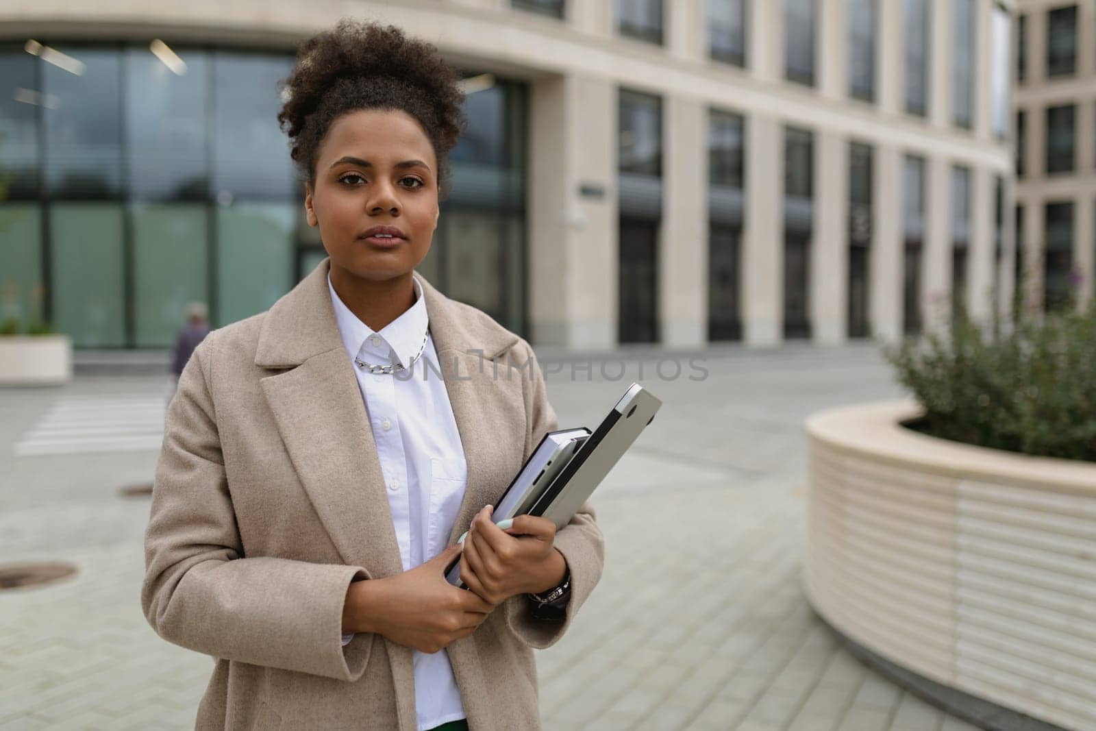 a woman of African American appearance, an official stands against the background of an administrative building with a laptop and documents in her hands.