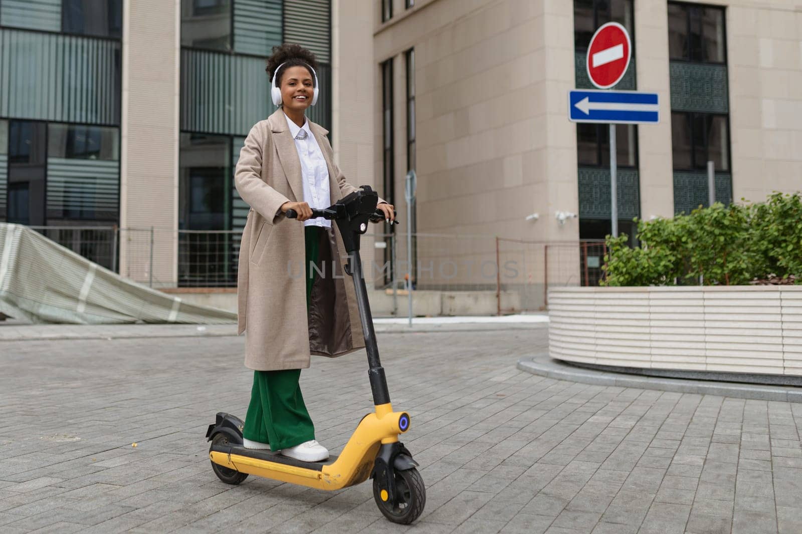stylish african american woman rides an electric scooter against the backdrop of an office building.