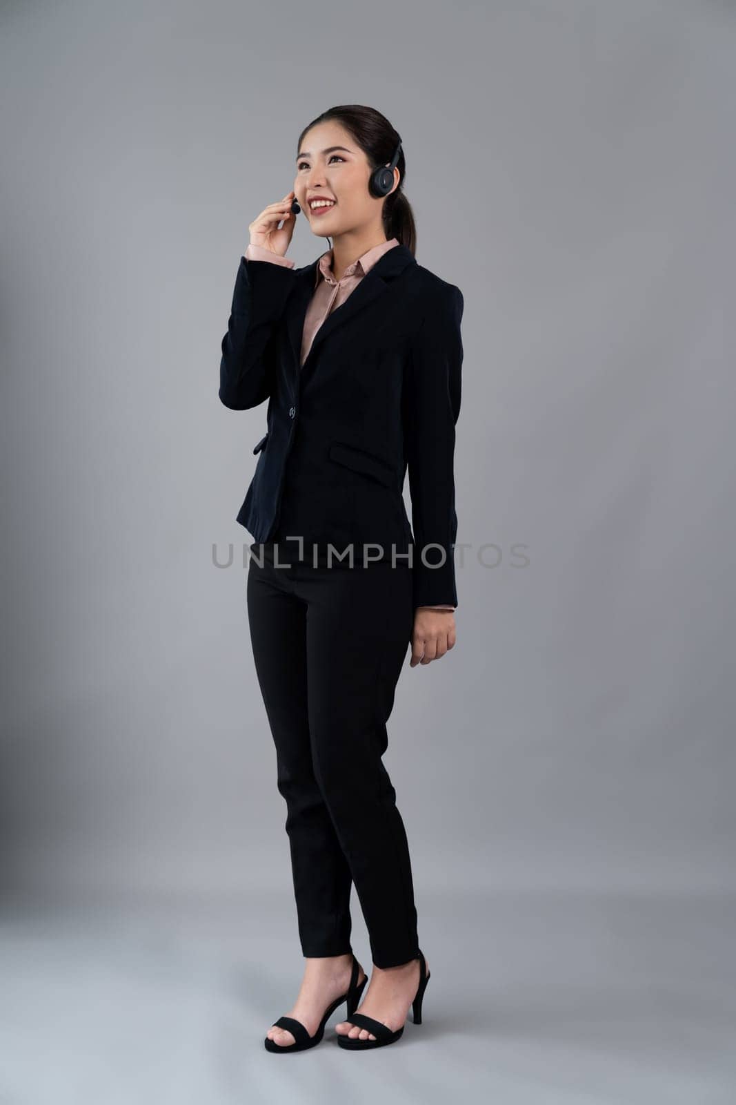 Attractive full body asian female call center operator with happy face advertises job opportunity on empty space, wearing formal suit and headset on customizable isolated background. Enthusiastic