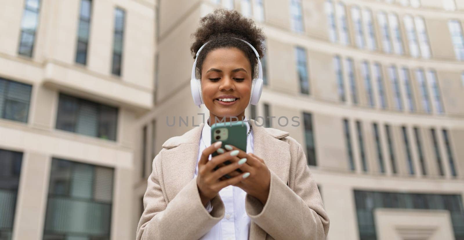 cheerful charismatic african american woman listening to music in headphones with a mobile phone in her hands outside by TRMK