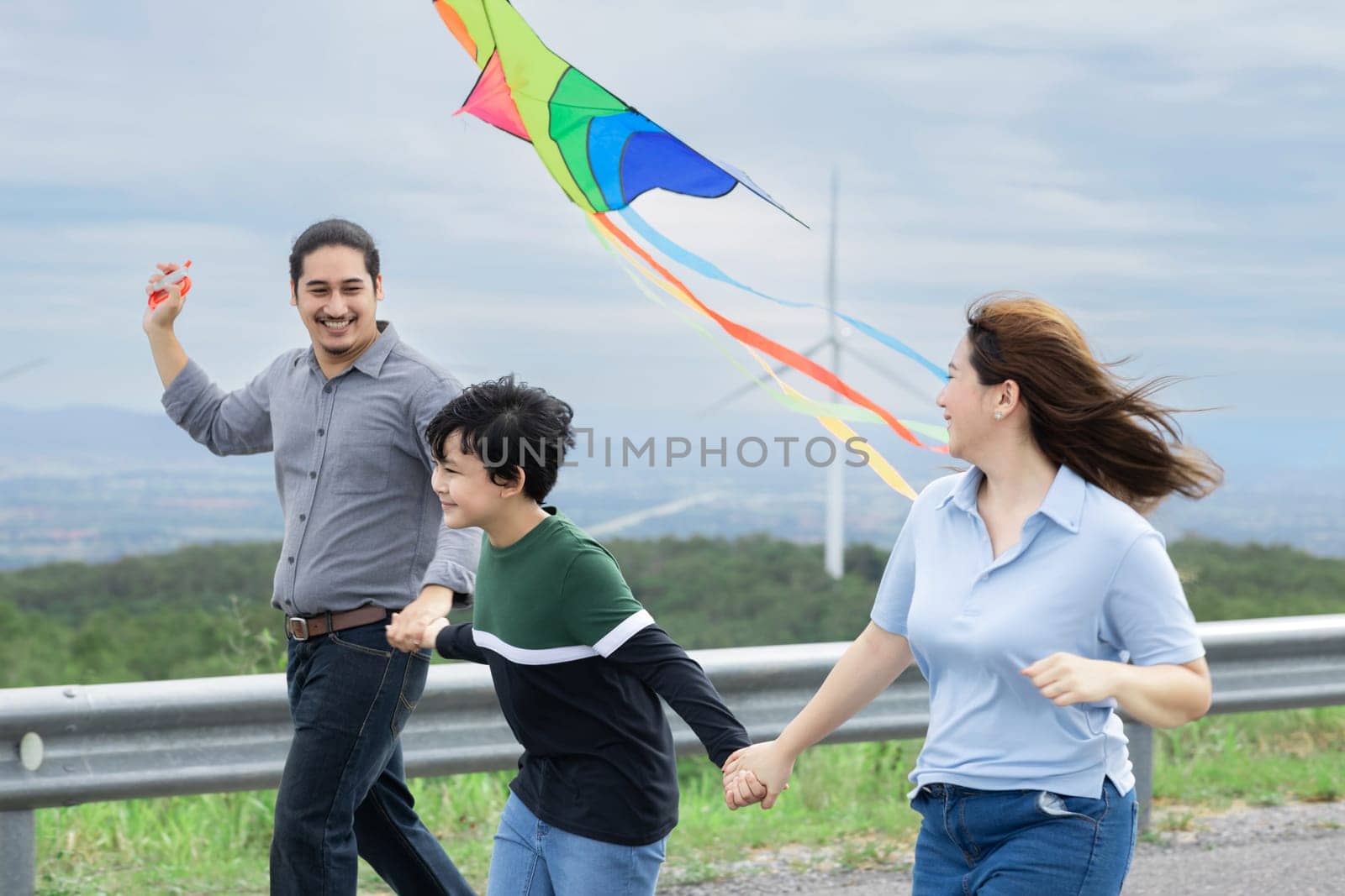 Progressive happy carefree family vacation concept. Young parents mother father and son run along and flying kite together on road with natural scenic on mountain and wind turbine background.