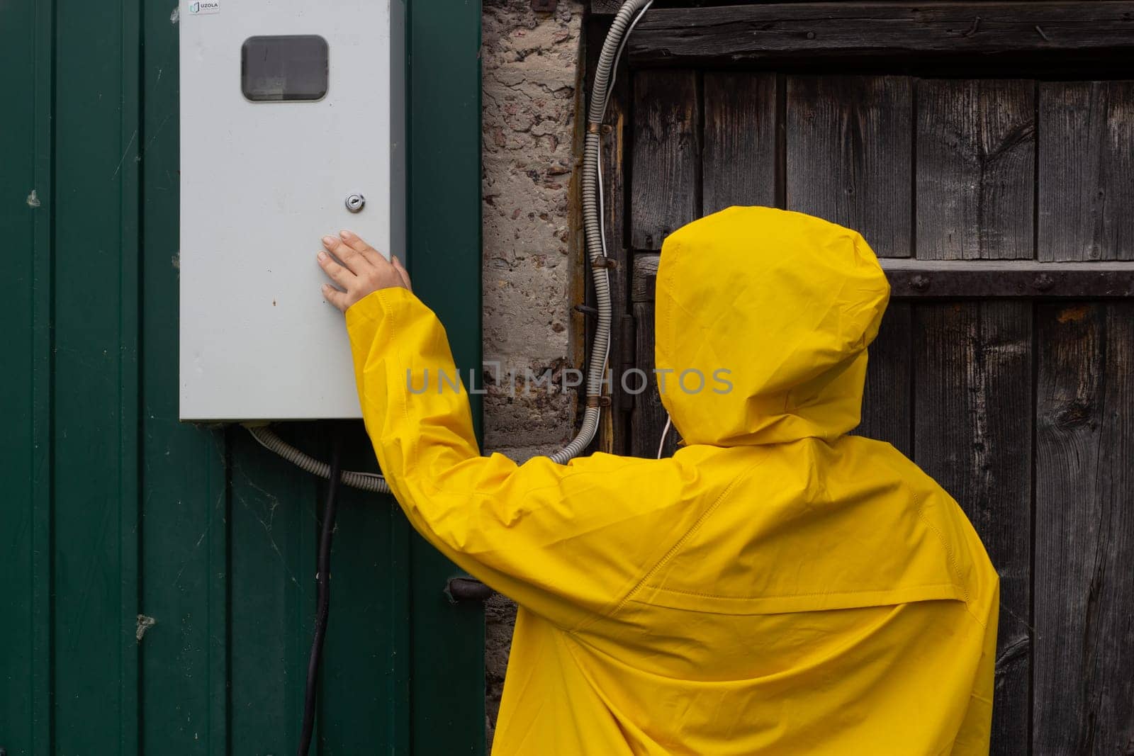 A woman in a yellow raincoat looks into a transformer box. Electricity and wires. View from the back.