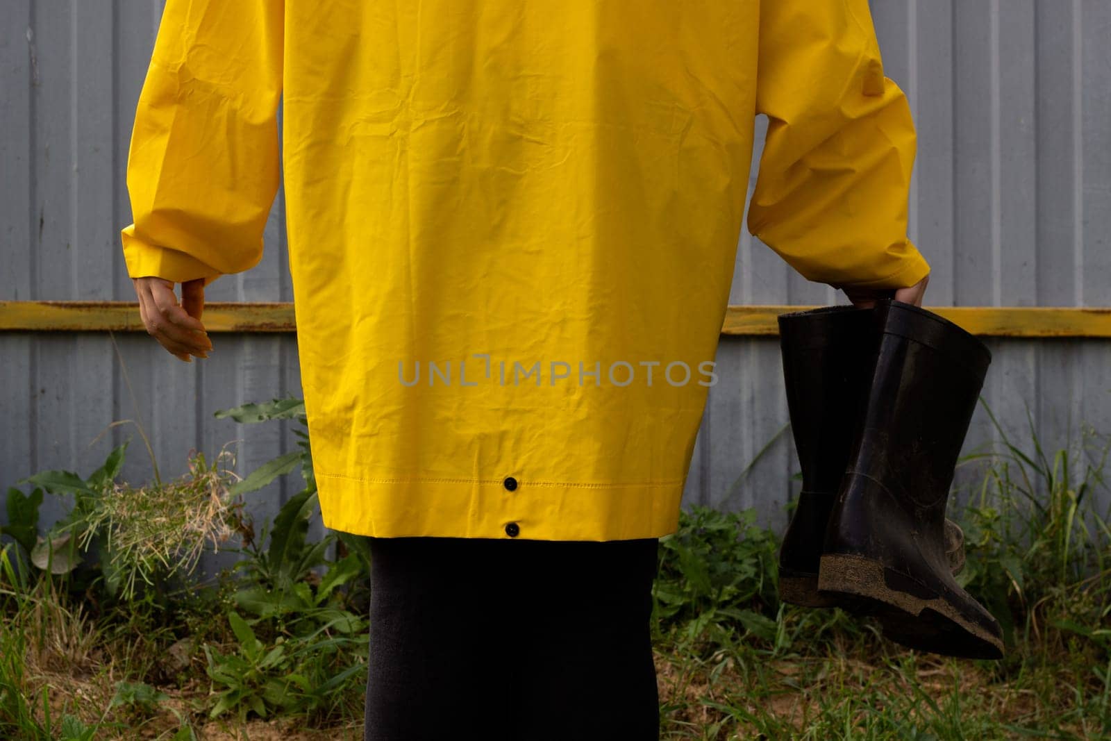 A woman in a yellow raincoat holds black rubber boots in her hand. Against the background of a gray metal fence.