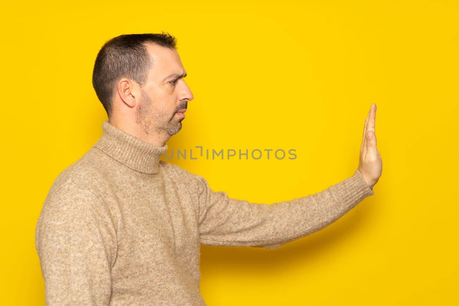 Rejection, denial concept. Side view of man wearing turtleneck making stop sign with outstretched hand, warning expression with negative gesture. Indoor studio shot isolated on yellow background.