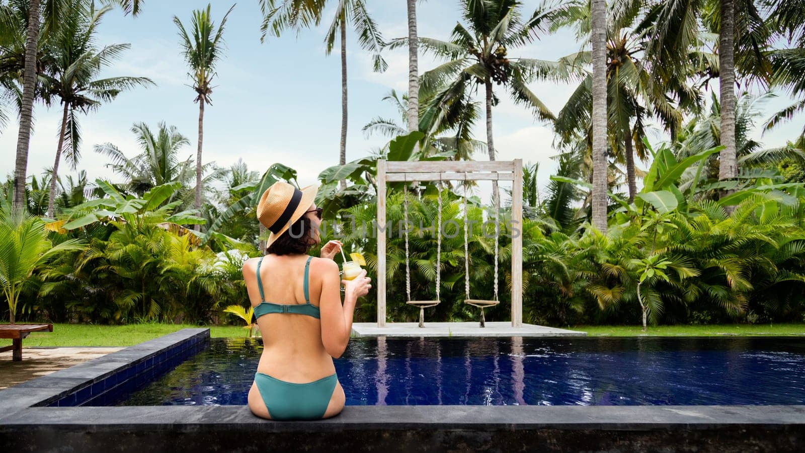 Rear view of young female relaxing in the swimming pool drinking pineapple juice on tropical vacation resort. Copy space. Panoramic image. Summer vacation concept.