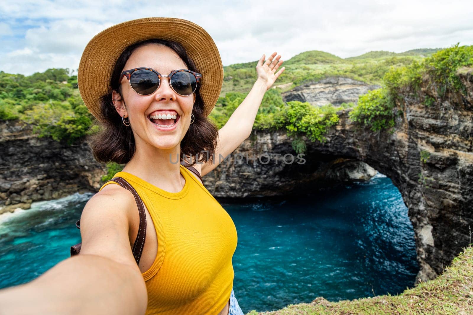 Young woman wearing hat and sunglasses taking selfie at Broken Beach, Nusa Penida, Bali, Indonesia. Female taking picture in summer vacation. Looking at camera. Copy space. Travel, freedom and happiness concept.