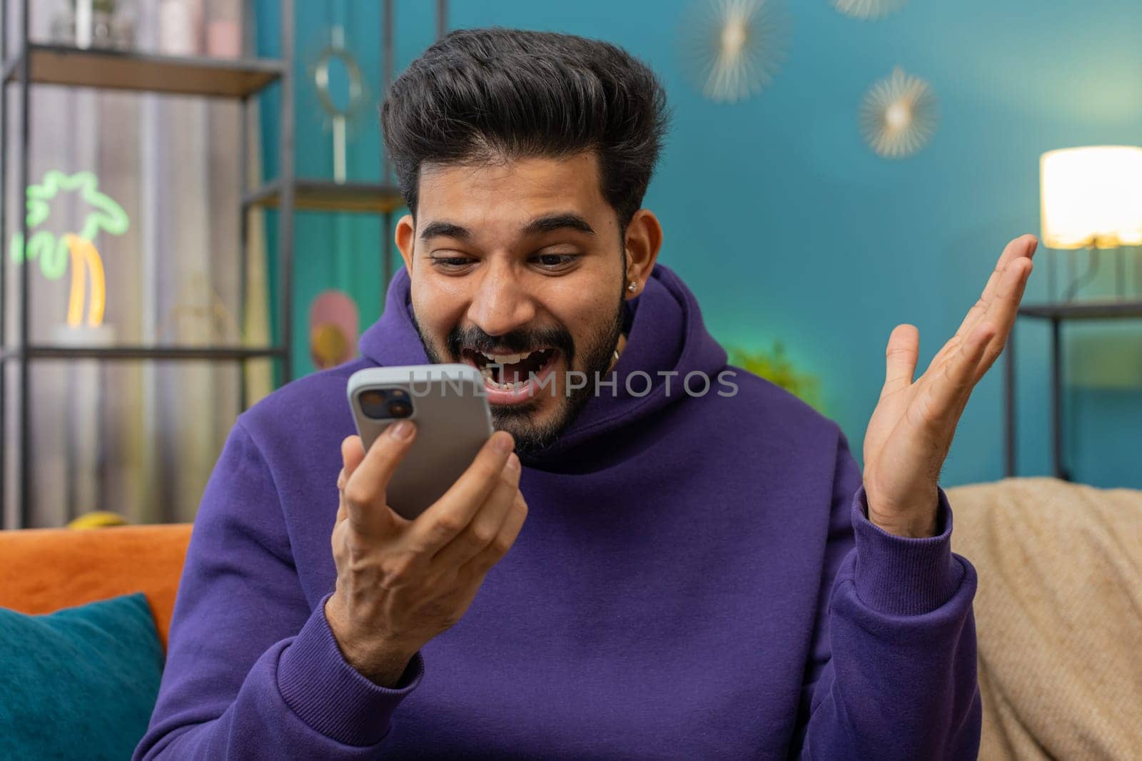 Happy indian man use mobile smartphone typing browsing say Wow yes found out great big win good news celebrate lottery jackpot doing winner gesture. Hindu young guy at home in room sitting on sofa