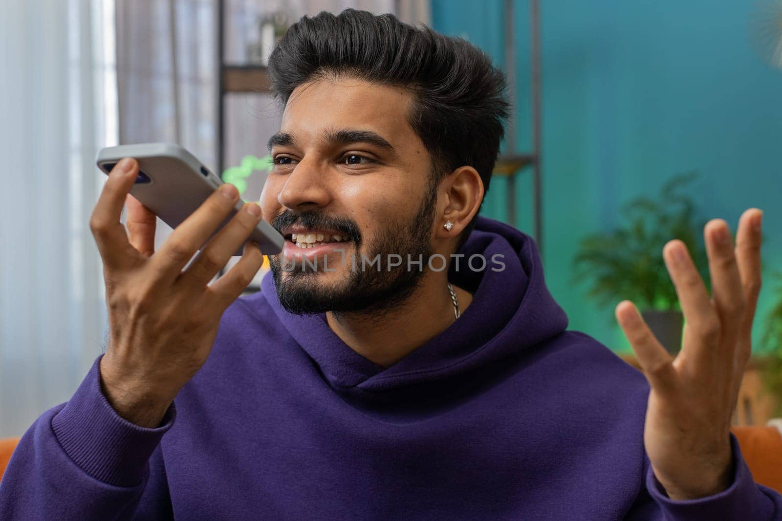 Happy excited indian man making online phone call conversation with friends. Portrait of hindu guy enjoying mobile loudspeaker talking chatting communication at home apartment living room on couch