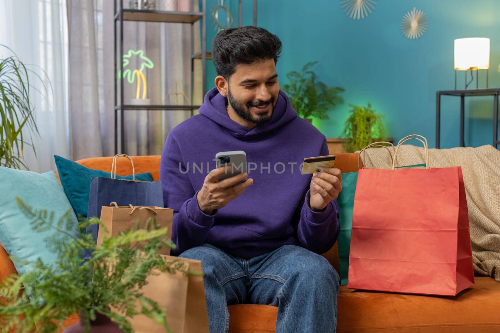Happy shopaholic consumer indian man sitting with shopping bags at modern home apartment making online order, payment with credit plastic bank card. Discount sale on internet. Young hindu guy on sofa