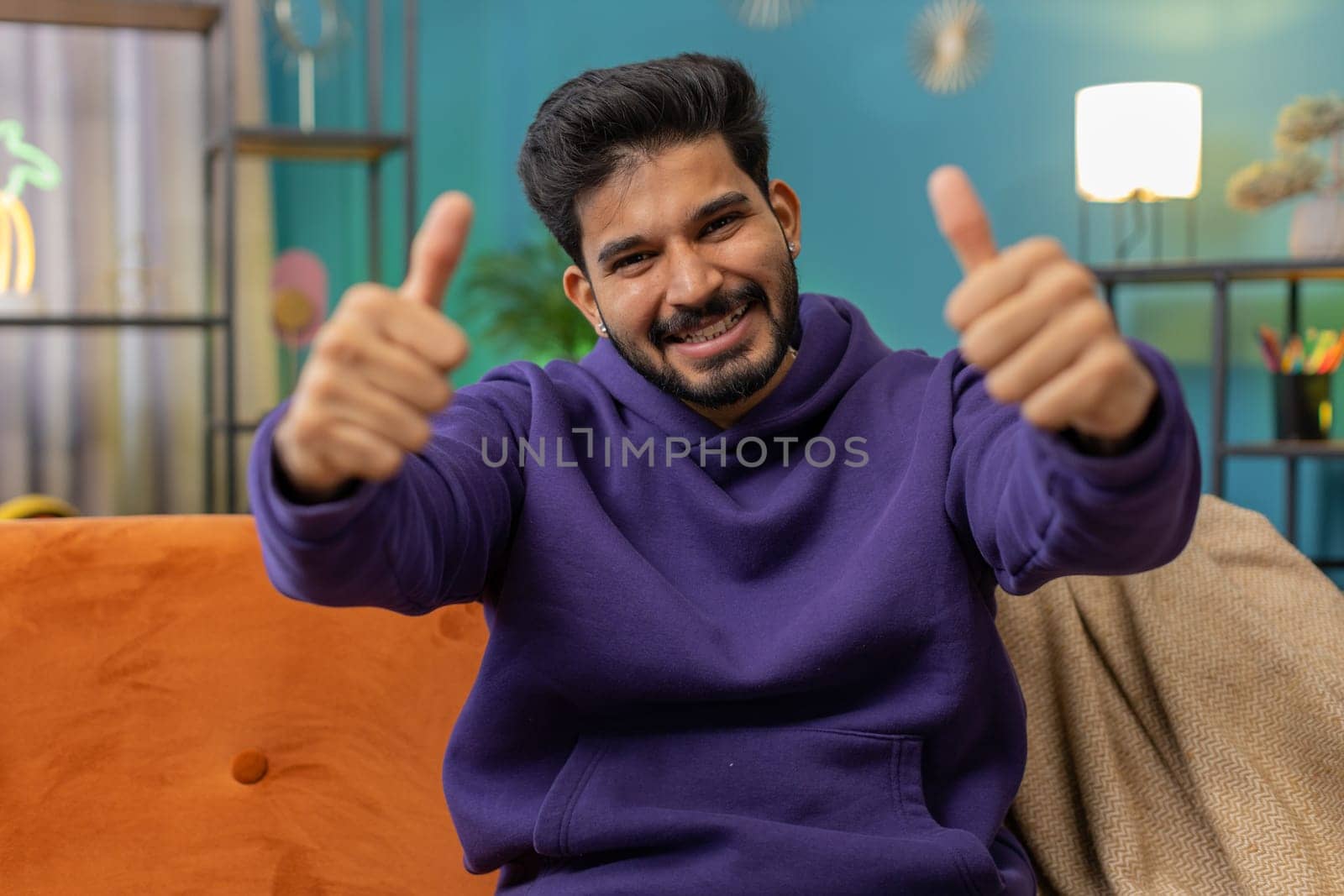 Like. Happy excited indian man looking approvingly at camera showing thumbs up, like sign positive something, good great news, positive feedback. Young hindu guy sitting on couch at home living room