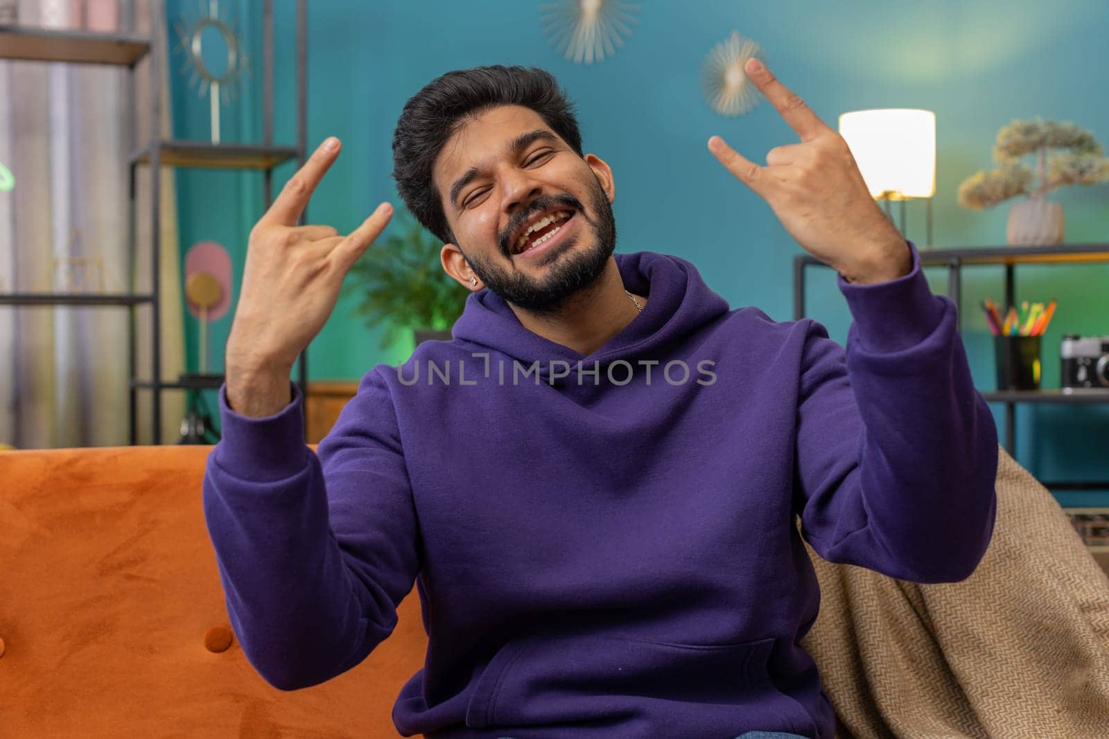 Overjoyed indian man showing rock n roll gesture by hands, cool sign, shouting yeah with crazy expression, dancing, emotionally rejoicing in success. Young hindu guy at home apartment room on couch