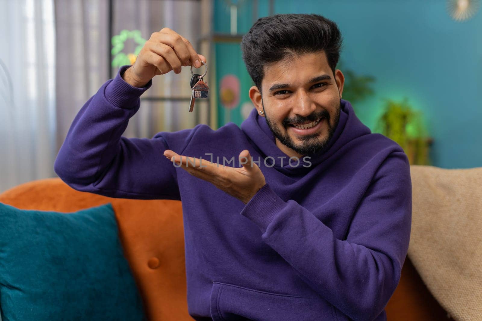 Indian man real estate agent lifting hand showing the keys of new home house apartment, buying or renting property, mortgage loan at modern home apartment indoors. Hindu guy in room sitting on sofa
