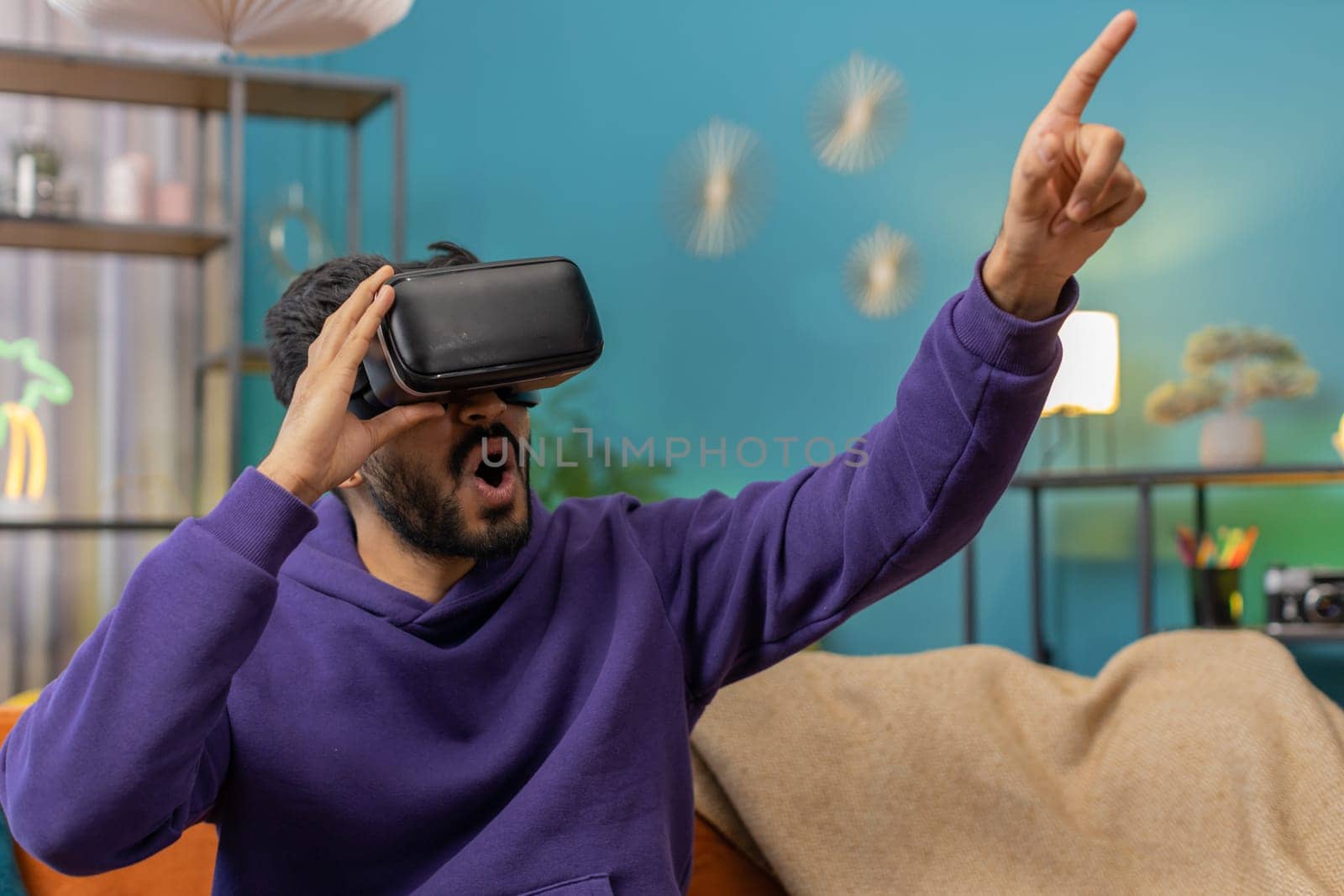 Indian man using virtual reality futuristic technology VR app headset helmet to play simulation 3D 360 video game, watching film movie at modern home apartment. Hindu guy in goggles sitting on couch