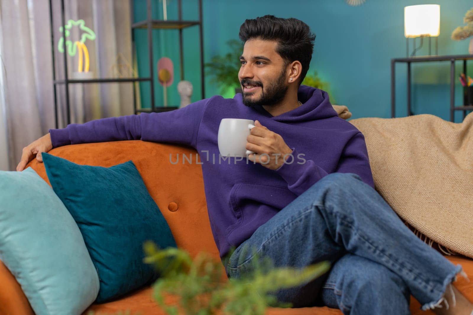Handsome smiling indian man drinking a cup of warm coffee or herbal tea at home living room apartment. Break time. Hindu guy enjoying comfortable relaxing sitting on couch in the morning. Lifestyles