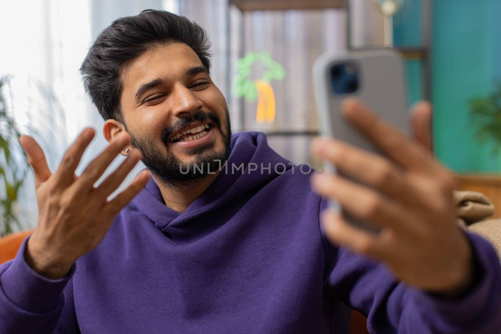 Happy indian man blogger taking selfie on smartphone, communicating video call online with social media subscribers followers, recording vlog stories. Young hindu guy at home apartment room on couch