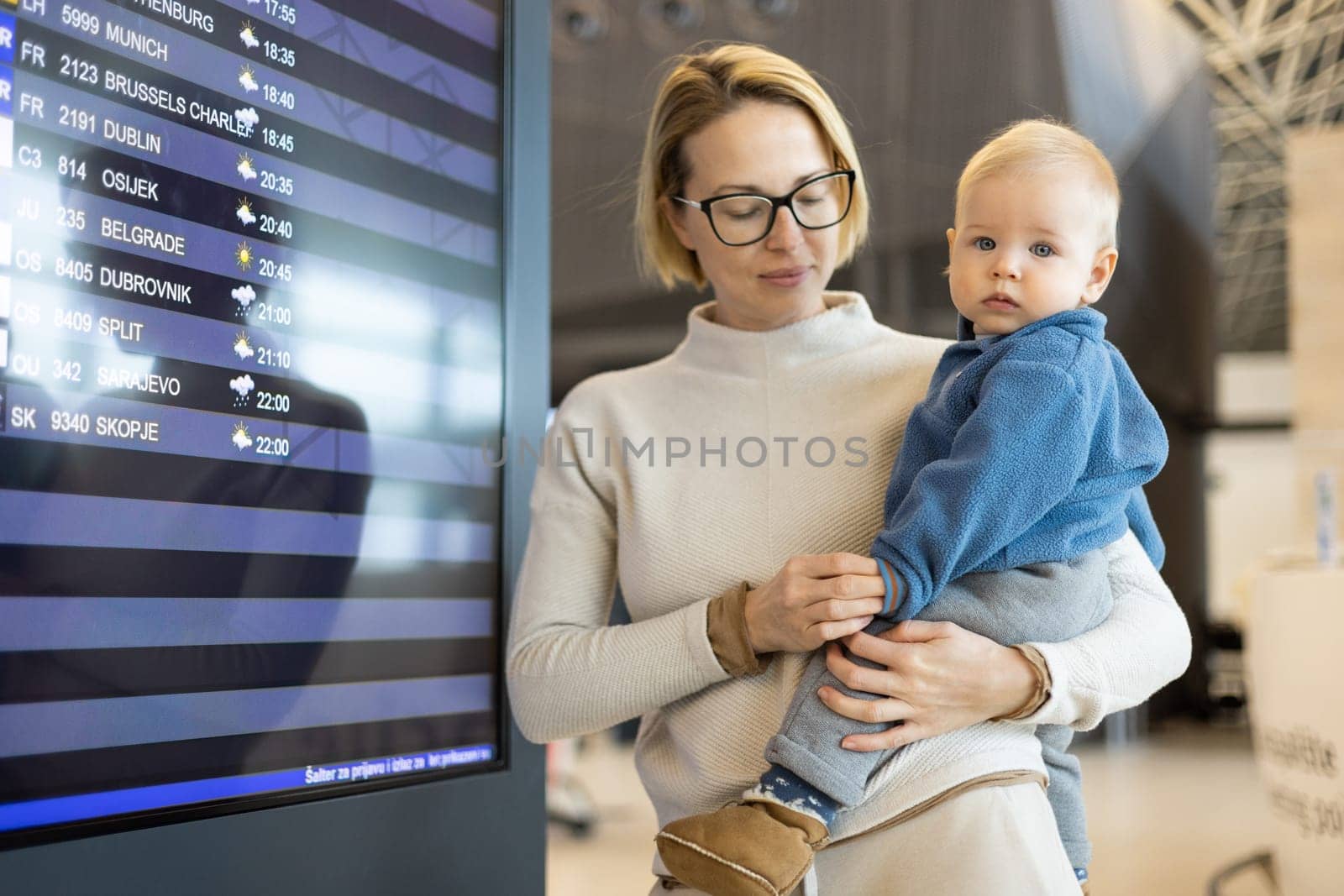 Mother traveling with child, holding his infant baby boy at airport terminal, checking flight schedule, waiting to board a plane. Travel with kids concept