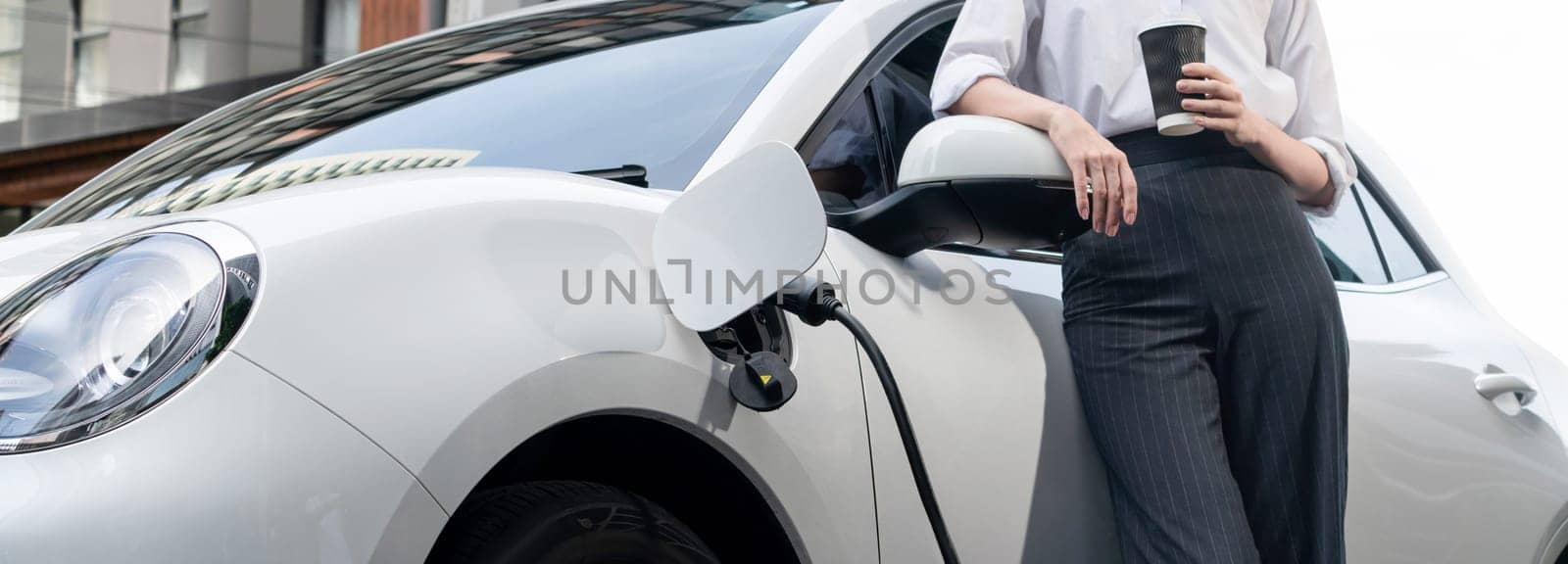 Closeup progressive suit-clad businesswoman with her electric vehicle recharge her car on public charging station in modern city with power cable plug and renewable energy-powered electric vehicle.