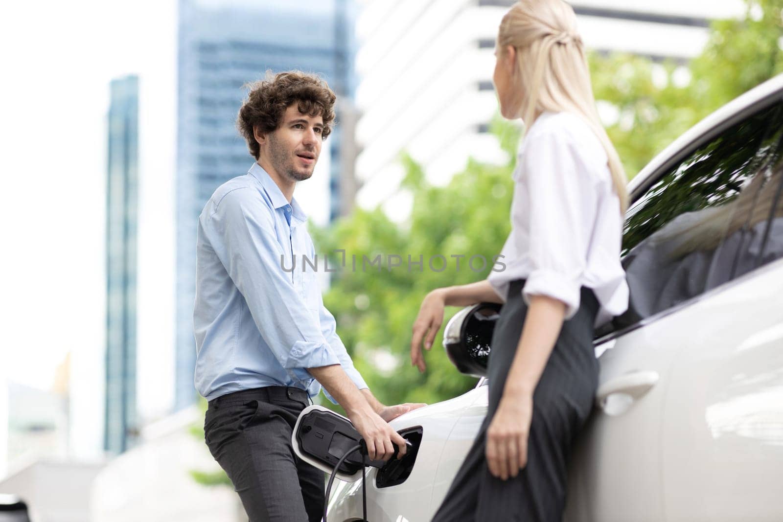 Progressive businessman and businesswoman with electric car connected to charging station before driving around city center. Eco friendly rechargeable car powered by alternative clean energy.