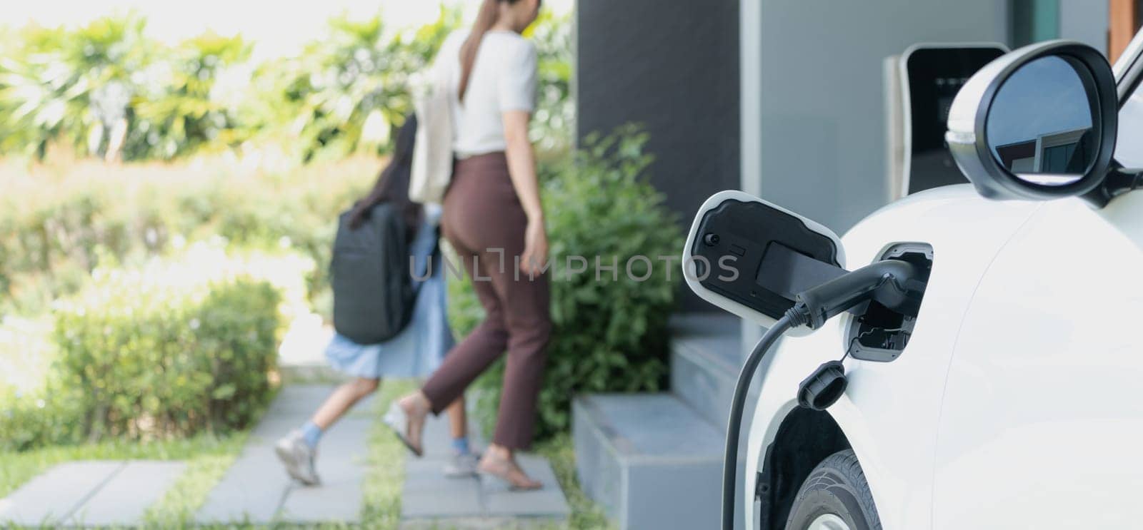 Focus EV car charging at home charging point with blurred background of family. by biancoblue