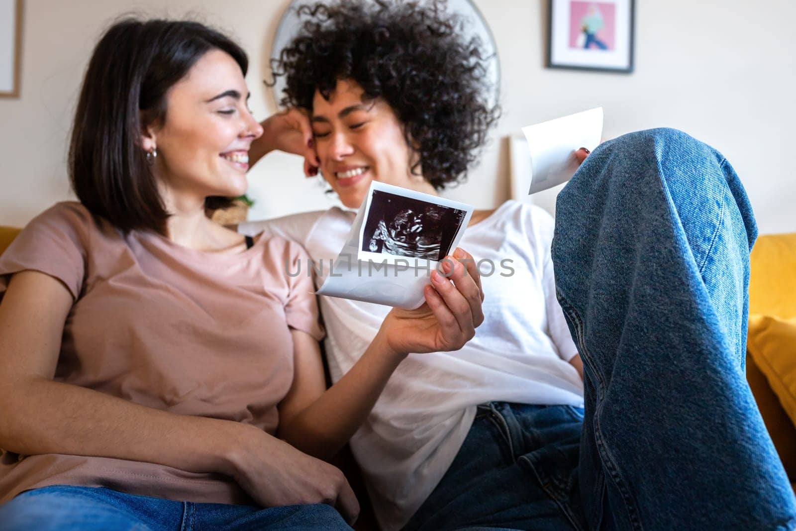 Bliss. Happy and joyful multiracial lesbian pregnant couple looking baby ultrasounds at home relaxing on the sofa. LGTBQ love and family concept.