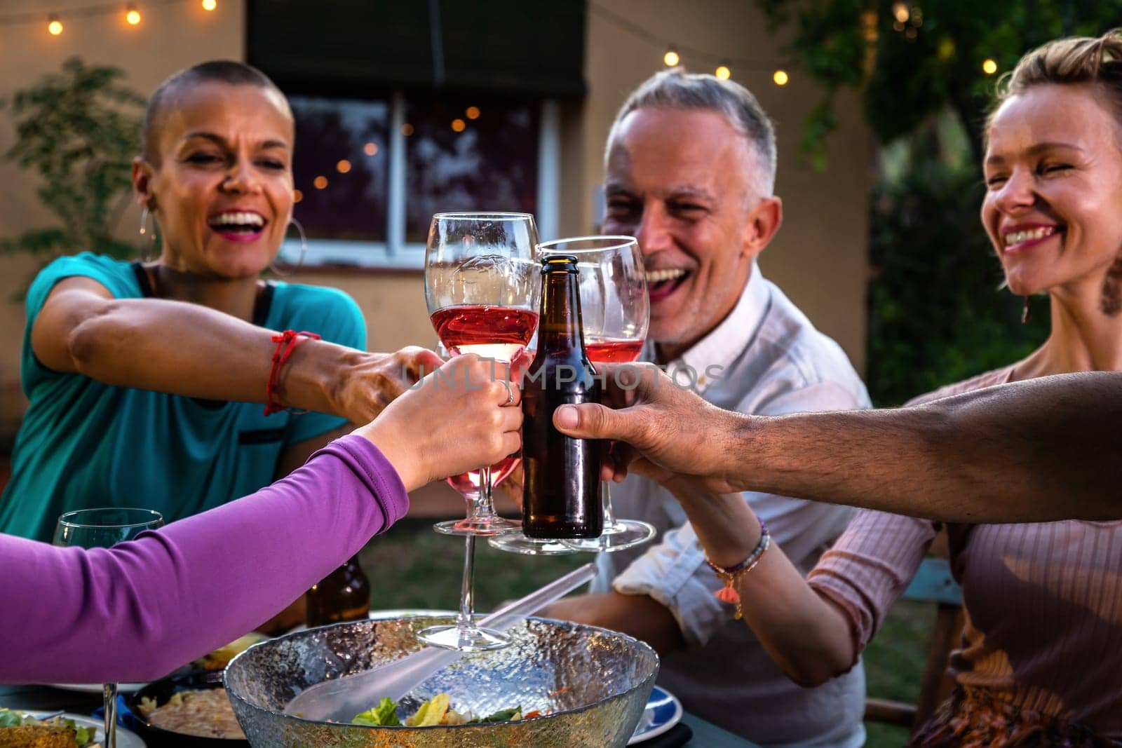 Close-up of smiling, happy friends having fun, toasting with wine and beer during garden dinner party at night. Lifestyle concept.