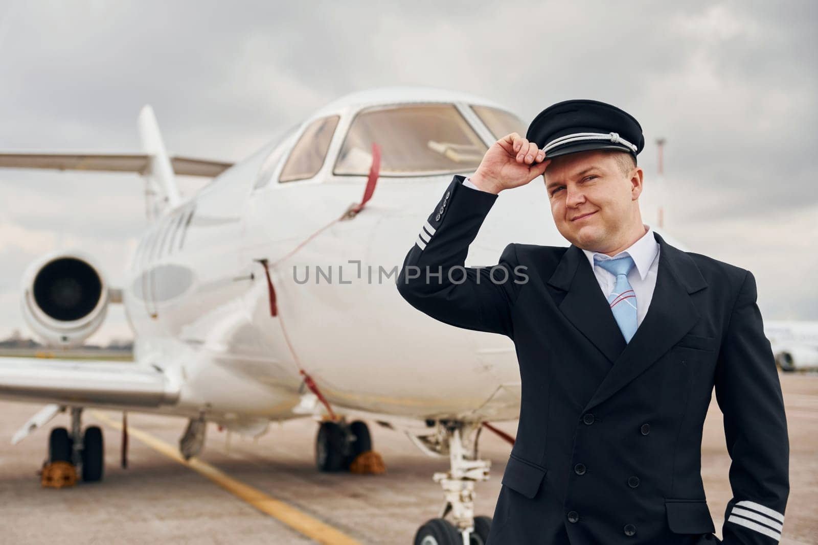 Posing for a camera. Experienced pilot in uniform standing outside near plane by Standret