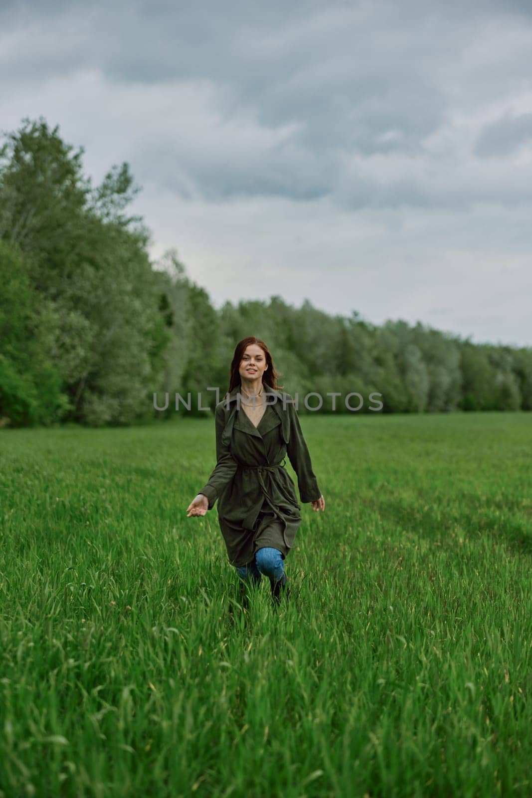 a beautiful woman in a long raincoat runs across a field in high grass in spring in cloudy weather. High quality photo