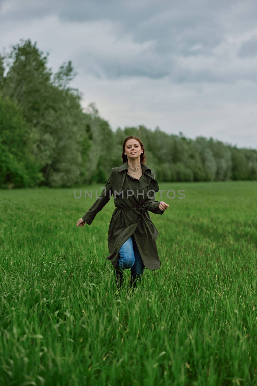 a beautiful woman in a long raincoat runs across a field in high grass in spring in cloudy weather. High quality photo