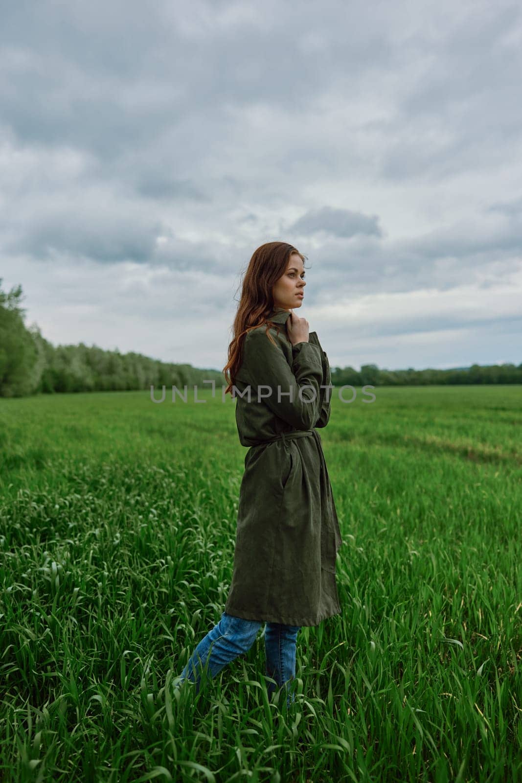 a beautiful woman in a dark coat stands in a green field in the spring in rainy weather. High quality photo