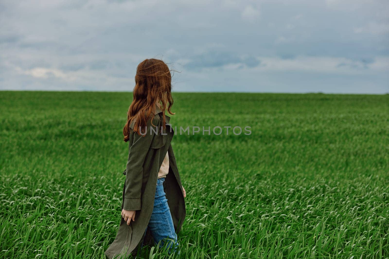 a red-haired woman in a long raincoat stands in a green field in cloudy weather in a strong wind by Vichizh