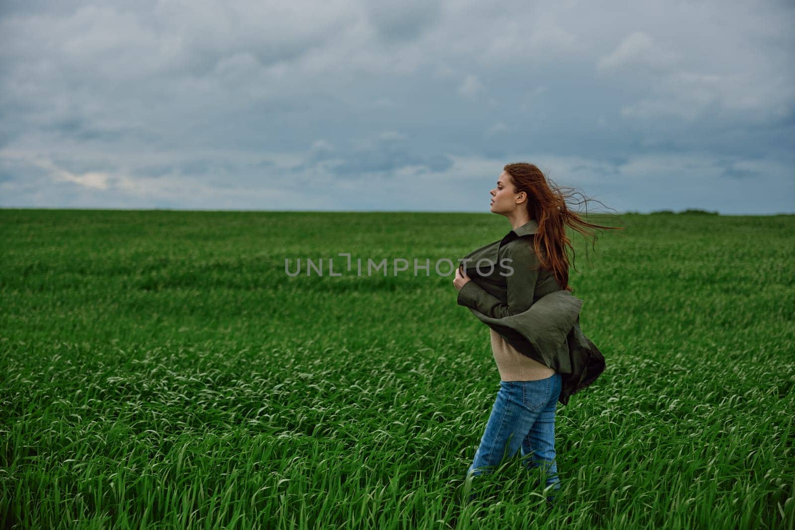 a red-haired woman stands in a green field in rainy, cold weather, holding a raincoat in the wind by Vichizh