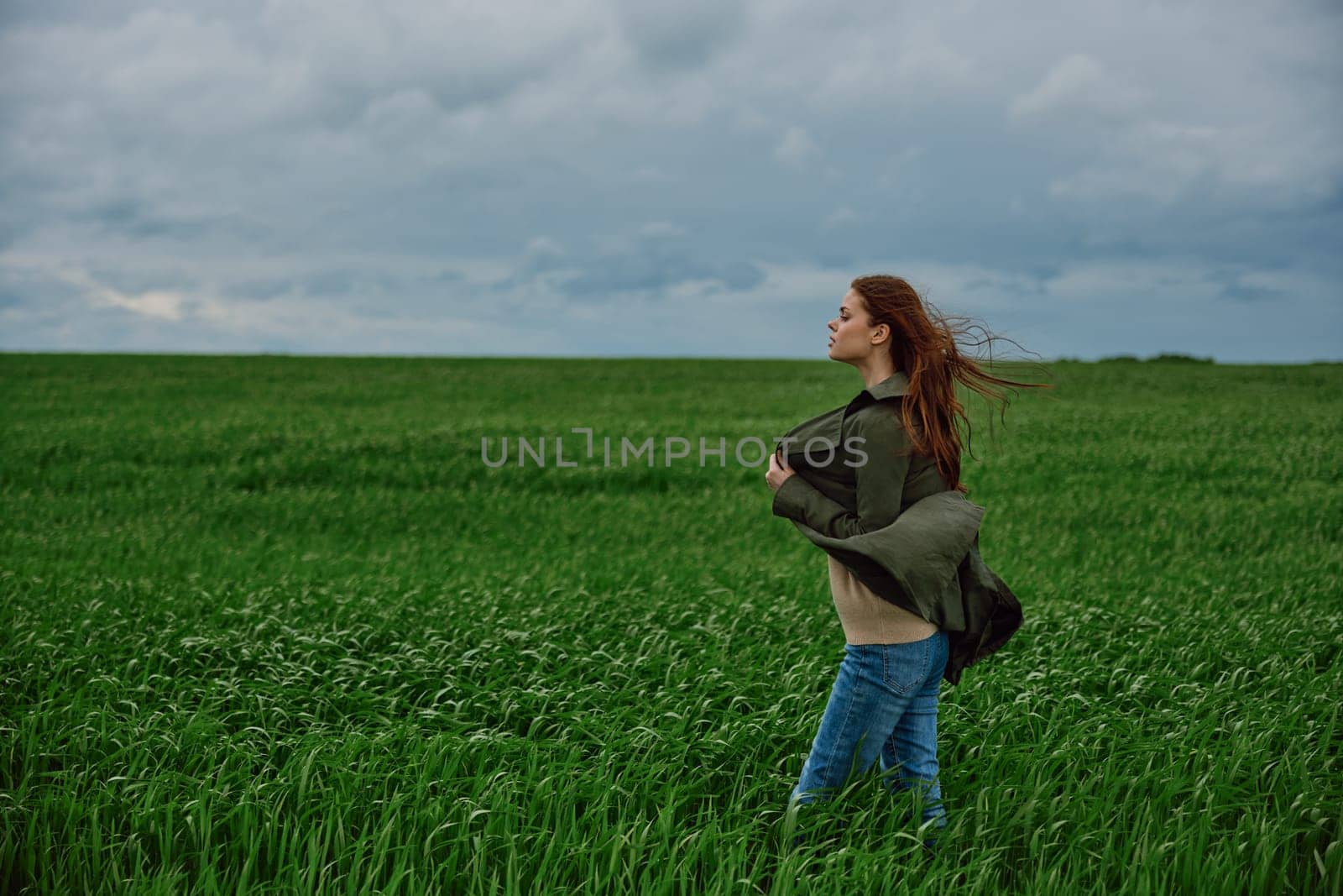 a red-haired woman stands in a green field in rainy, cold weather, holding a raincoat in the wind. High quality photo