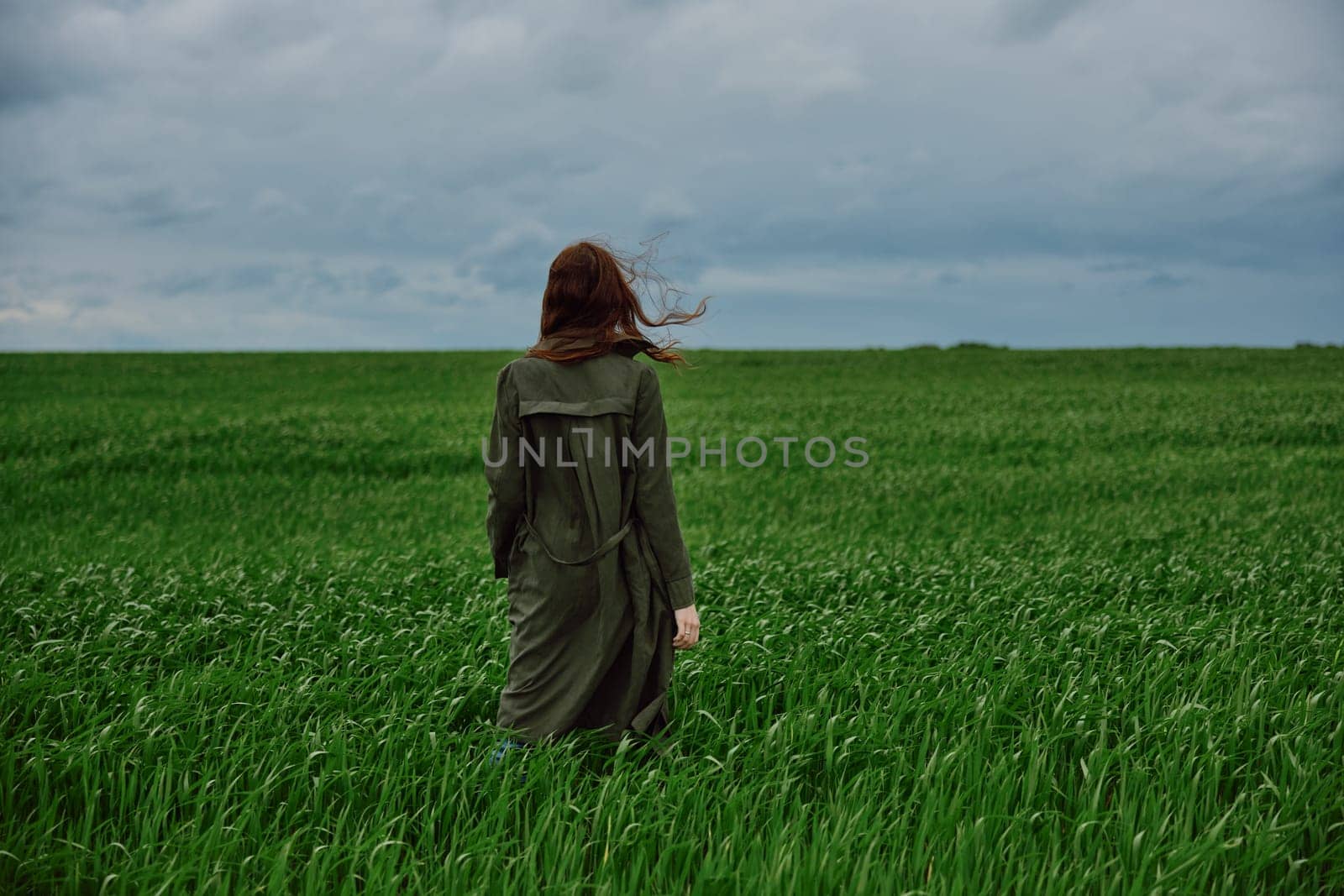 a woman in a long raincoat stands in a green field in windy weather with her back to the camera. High quality photo