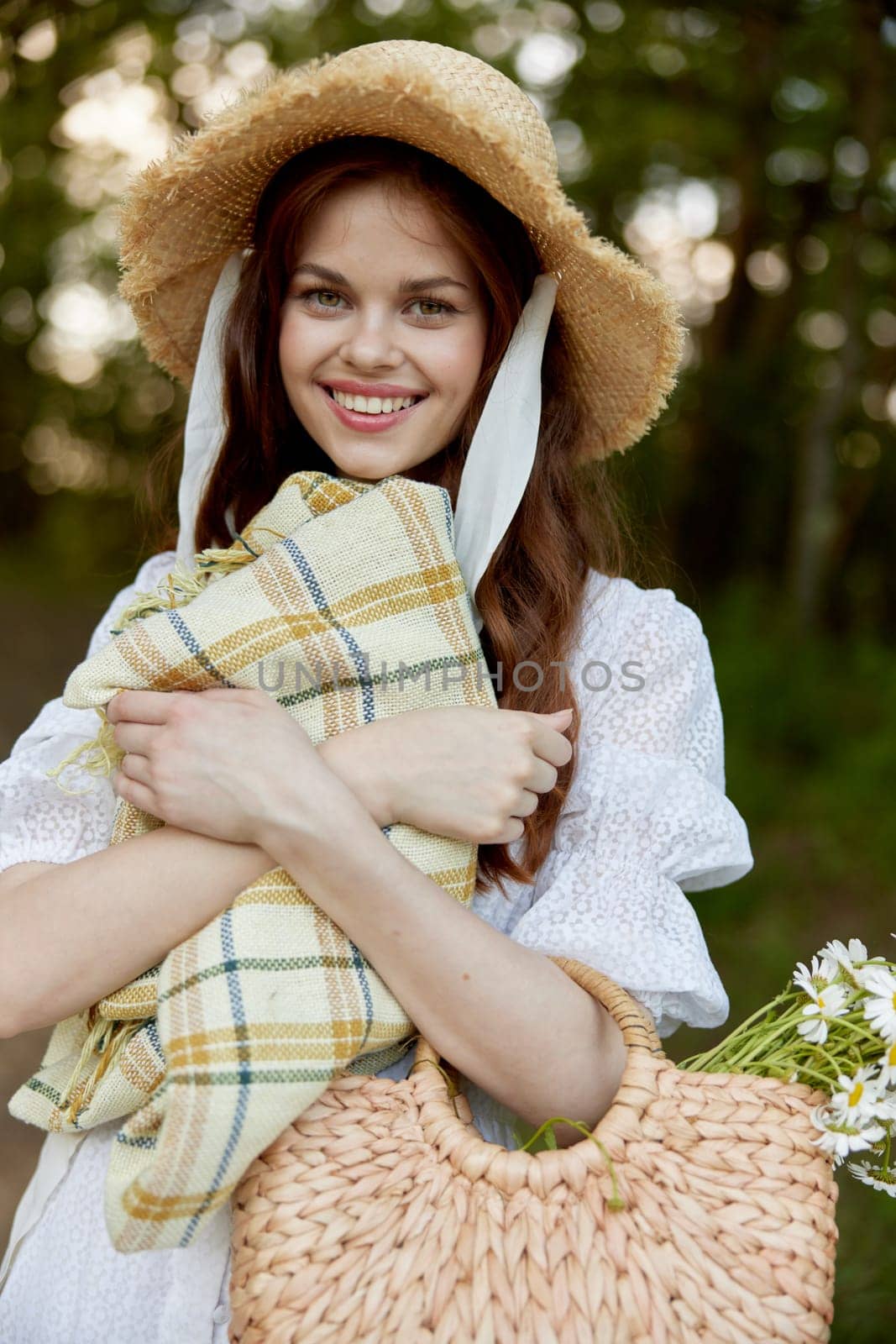portrait of a beautiful, happily smiling woman in a light dress, a plaid in her hands and with a wicker hat enjoys outdoor recreation by Vichizh