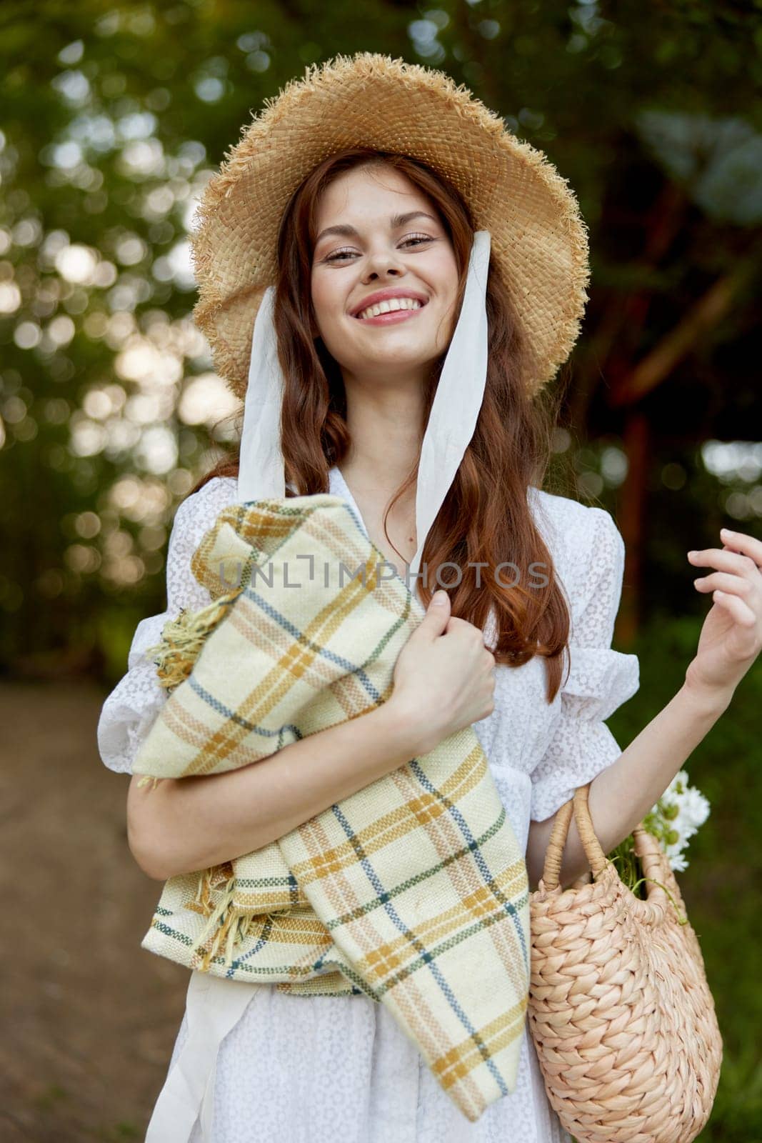 portrait of a beautiful, happily smiling woman in a light dress, a plaid in her hands and with a wicker hat enjoys outdoor recreation. High quality photo