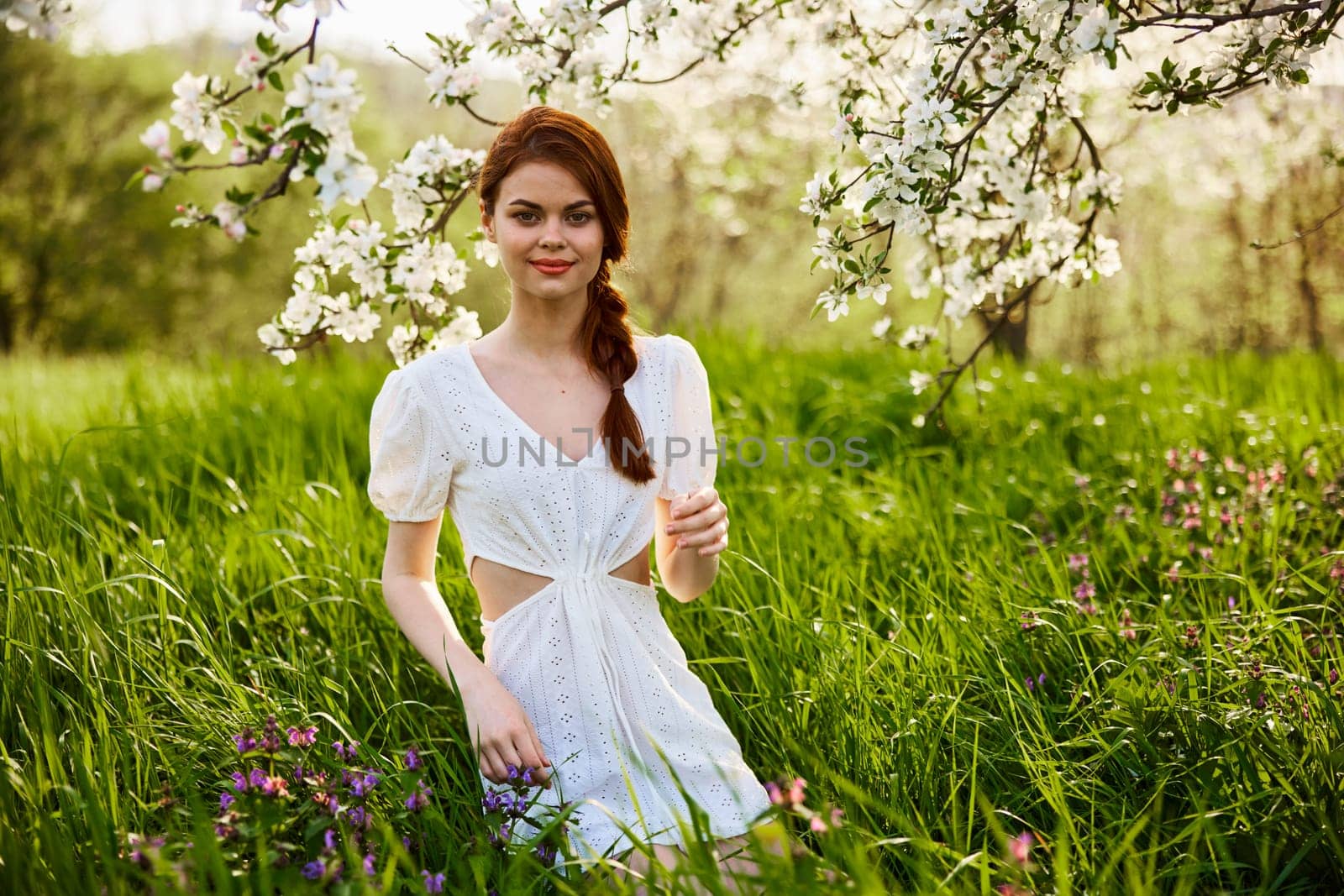 a young woman in a light summer dress stands near a tree with flowers and smiles looking at the camera by Vichizh