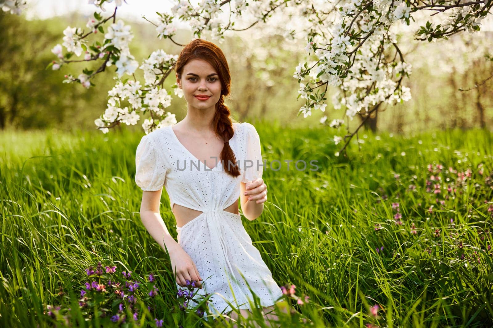 a young woman in a light summer dress stands near a tree with flowers and smiles looking at the camera. High quality photo