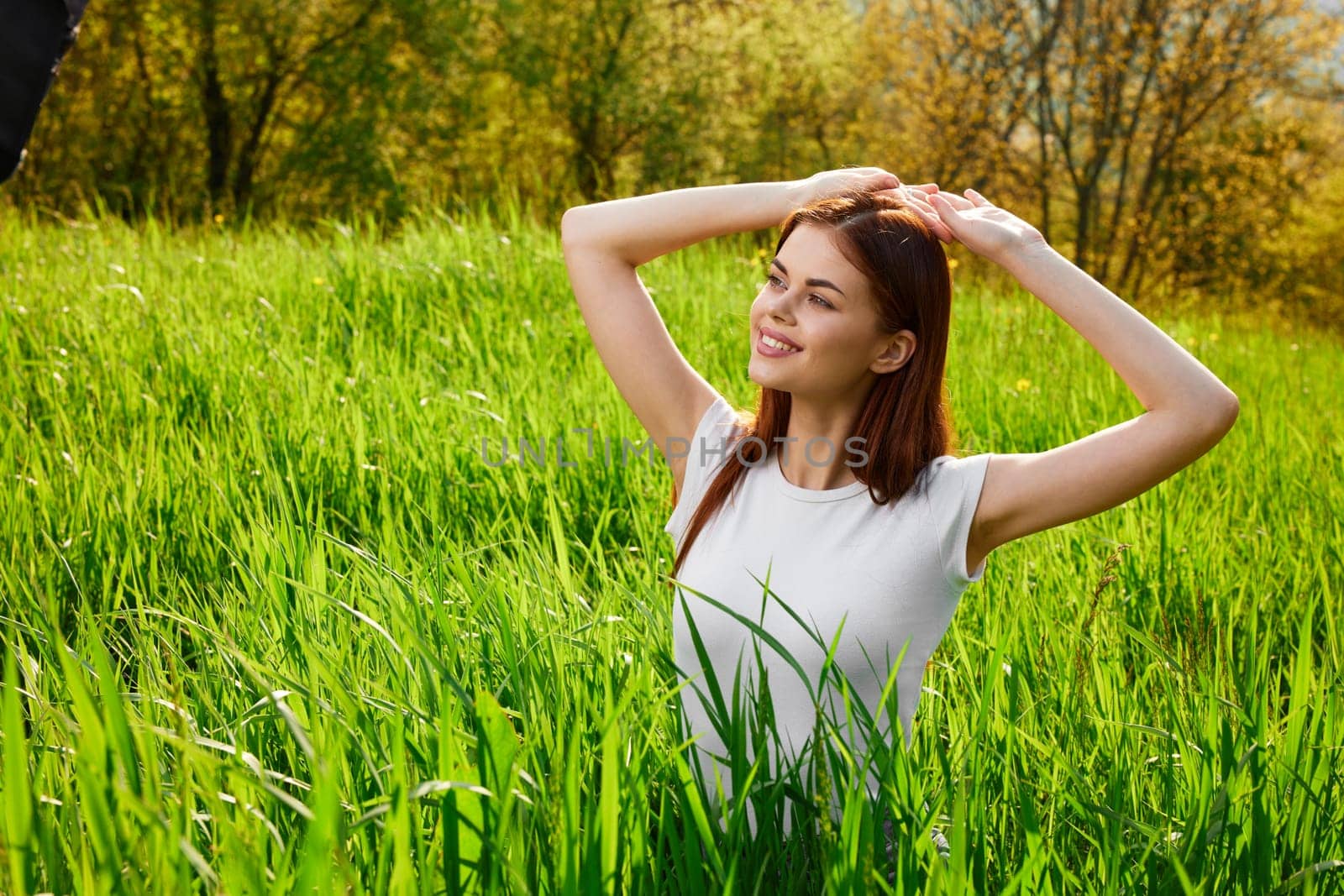portrait of a red-haired woman in a white T-shirt walking in the grass to the waist and holding her hands near her head. High quality photo