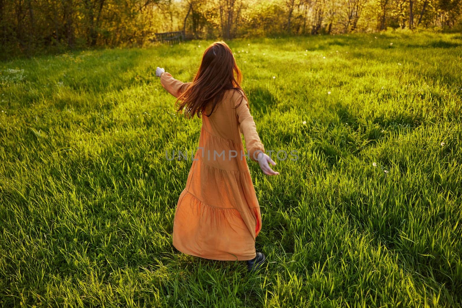 an attractive, slender, red-haired woman stands in a wide, green field during sunset in a long orange dress enjoying unity with nature and relaxation raising her arms to the sides while standing with her back to the camera. High quality photo