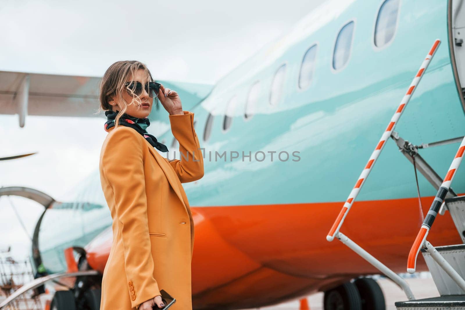 Passenger woman that is in yellow clothes, sunglasses and with luggage is outdoors near plane by Standret