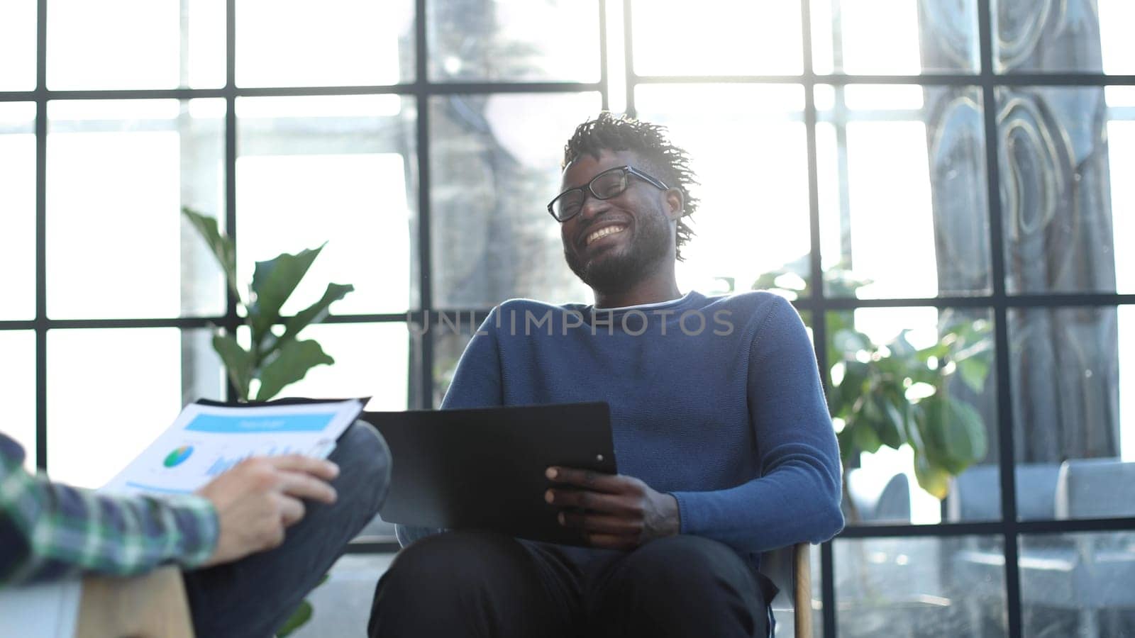 black man with a folder in his hands laughs while sitting on a chair