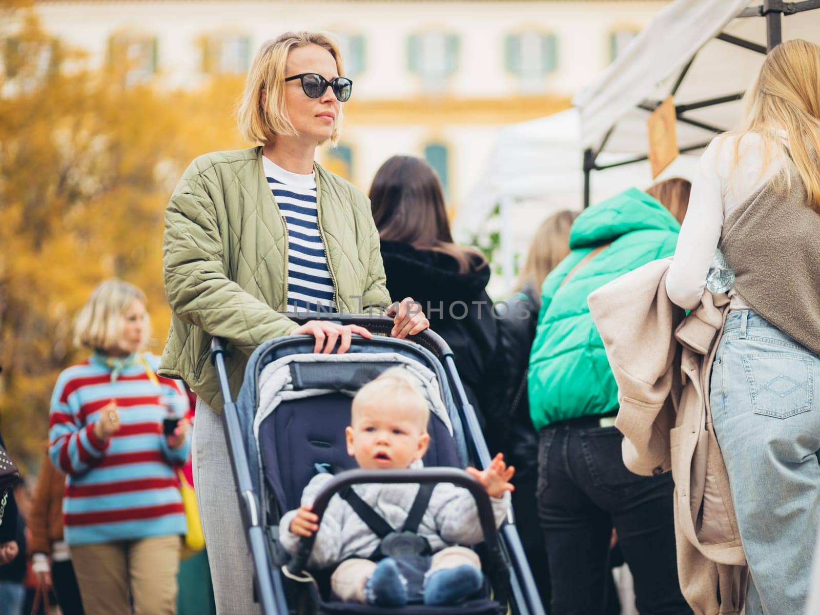 Mother waling and pushing his infant baby boy child in stroller in crowd of people wisiting sunday flea market in Malaga, Spain