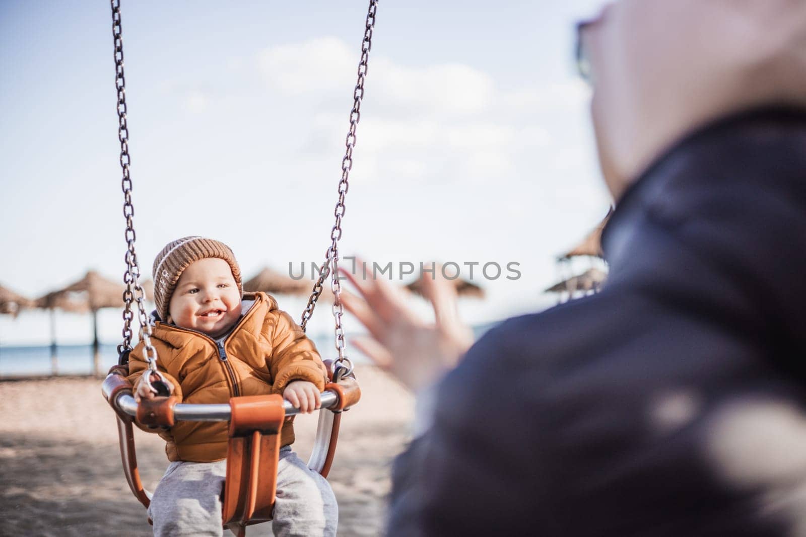 Mother pushing her cheerful infant baby boy child on a swing on sandy beach playground outdoors on nice sunny cold winter day in Malaga, Spain