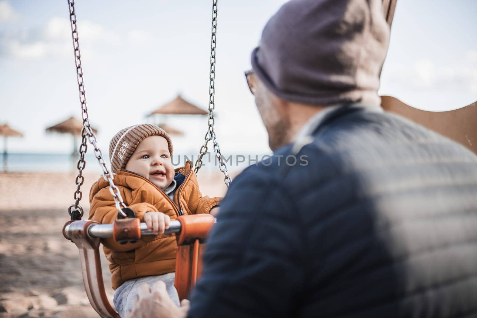 Father pushing hir cheerful infant baby boy child on a swing on sandy beach playground outdoors on nice sunny cold winter day in Malaga, Spain. by kasto