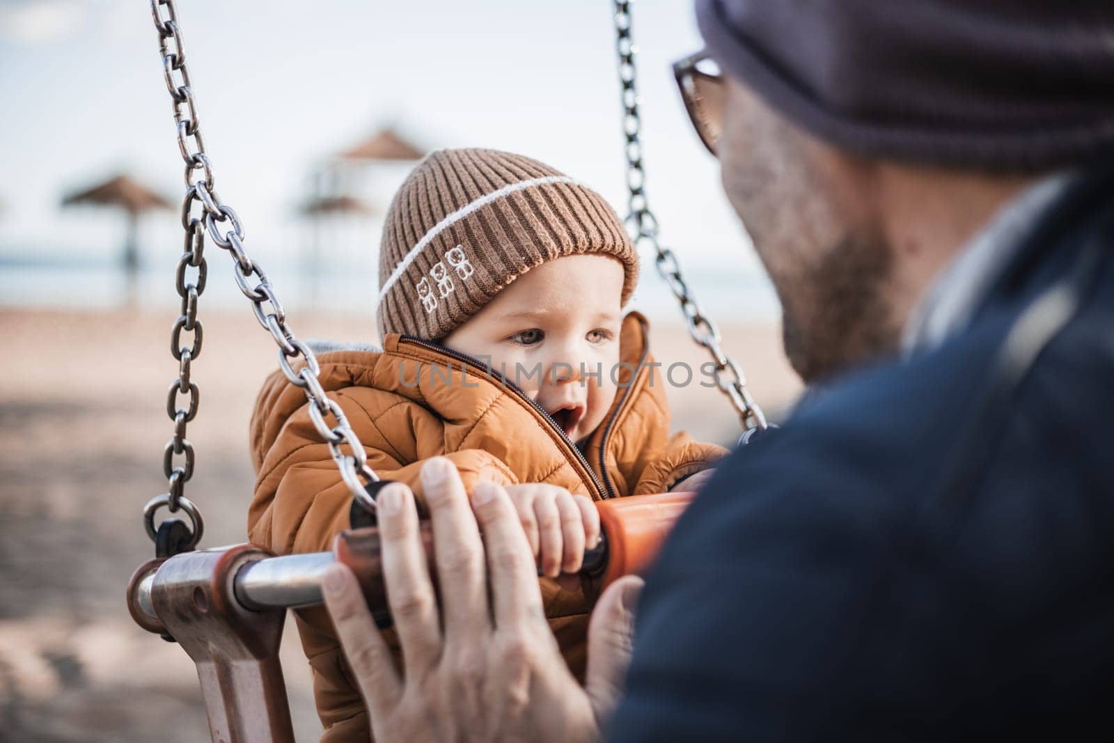 Father pushing hir cheerful infant baby boy child on a swing on sandy beach playground outdoors on nice sunny cold winter day in Malaga, Spain. by kasto