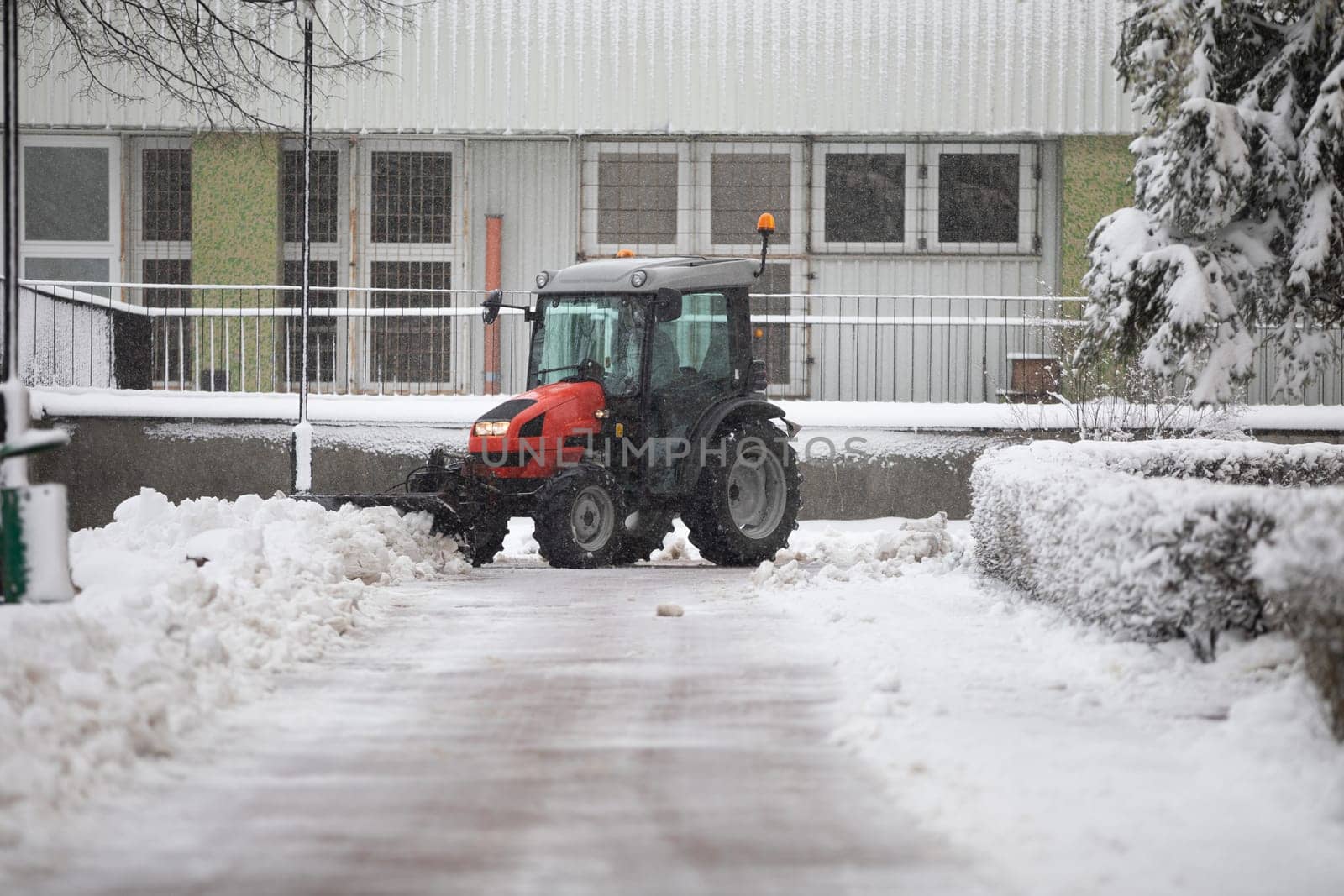 A red vehicle clears snow from the building's driveway. Removing fresh snow from the parking lot in front of the block. Winter in the city. Intense snowfall makes it difficult to drive a car.